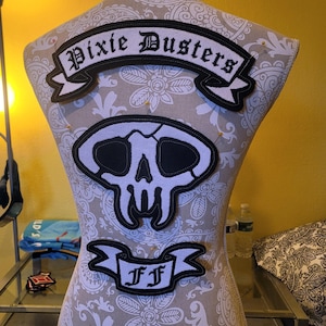 May include: A white mannequin with a floral pattern on its torso is wearing three black and white patches. The top patch is a banner with the text "Pixie Dustlers". The middle patch is a skull with a white background and black details. The bottom patch is a banner with the text "FF".