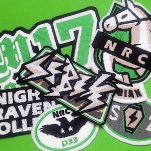 May include: A collection of embroidered patches featuring various designs, including a green and white patch with the text "NRC", a gray patch with the text "SZ", a green patch with the text "17", a white patch with the text "NIGHT RAVEN COLLEGE", and a gray patch with the text "RAZ".