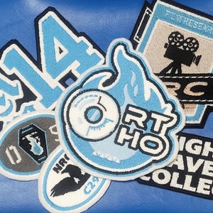 Może przedstawiać: Zbiór haftowanych naszywek z różnymi wzorami, w tym numerem "14", płomieniem z tekstem "ORTHO", płytą z białym środkiem, kamerą filmową z tekstem "FILM RESEARCH", czarno-białym ptakiem z tekstem "NIR C29" oraz czarną naszywką z tekstem "NIGHT HAVEN COLLEGE".