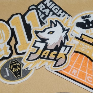 Puede incluir: Una colección de parches bordados con varios diseños, incluyendo una cabeza de lobo, el número "1", un logotipo de pista y campo, una calavera y huesos cruzados, y un logotipo con el texto "NRC 811".