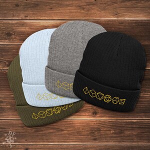 May include: Four ribbed knit beanies in olive green, light blue, gray, and black. Each beanie features a gold embroidered dice design along the folded brim. The hats are displayed on a wooden surface.