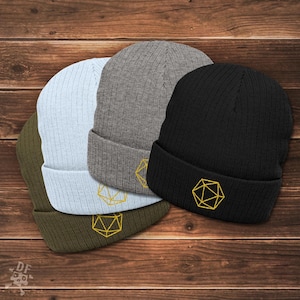 May include: Four ribbed knit beanies in olive green, light blue, gray, and black. Each beanie features a gold geometric design embroidered on the folded brim. The beanies are displayed on a wooden surface.