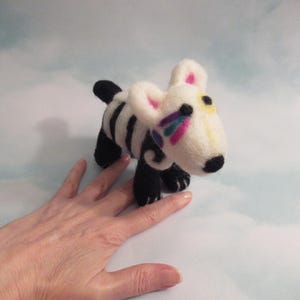 May include: A handmade felt dog figurine with a white body, black stripes, and black paws. The dog has a black nose, yellow and pink accents, and colorful decorative details around the eye. The figurine is approximately 4 inches long.