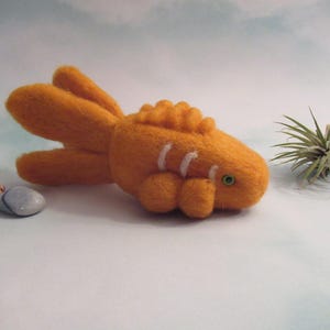 May include: A hand-felted orange goldfish with white stripes and a green eye. The goldfish is lying on a white surface.