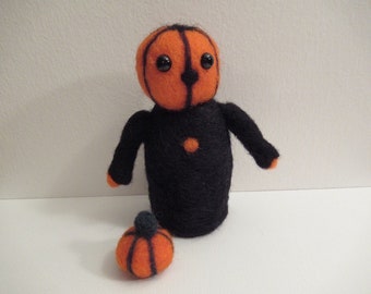 May include: A needle-felted doll with a round orange head featuring black vertical lines and two black bead eyes. The body is black with a small orange circular accent on the chest and orange tips on the arms. A small orange pumpkin sits nearby.