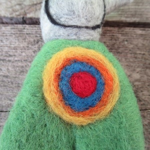 May include: A green felt toy with a colorful circular design on the back. The design features concentric circles of yellow, orange, blue, and red.