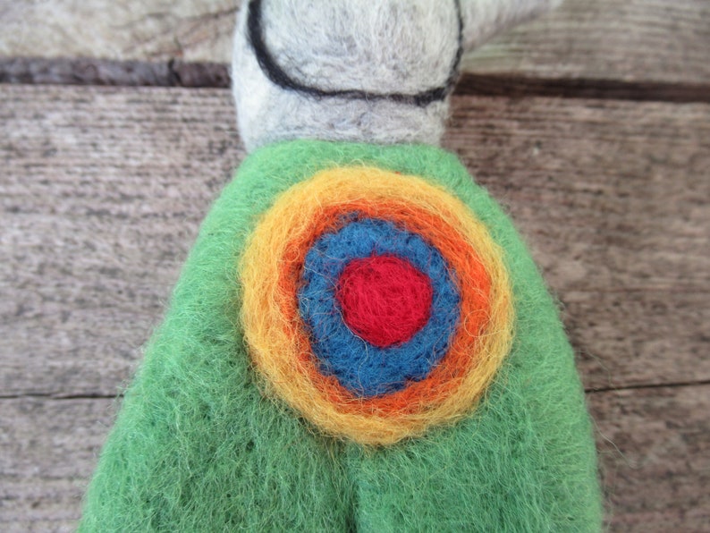 May include: A green felt toy with a colorful circular design on the back. The design features concentric circles of yellow, orange, blue, and red.