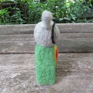 May include: A felt doll with a gray head, green body, and orange and yellow accents. The doll is standing on a wooden surface.