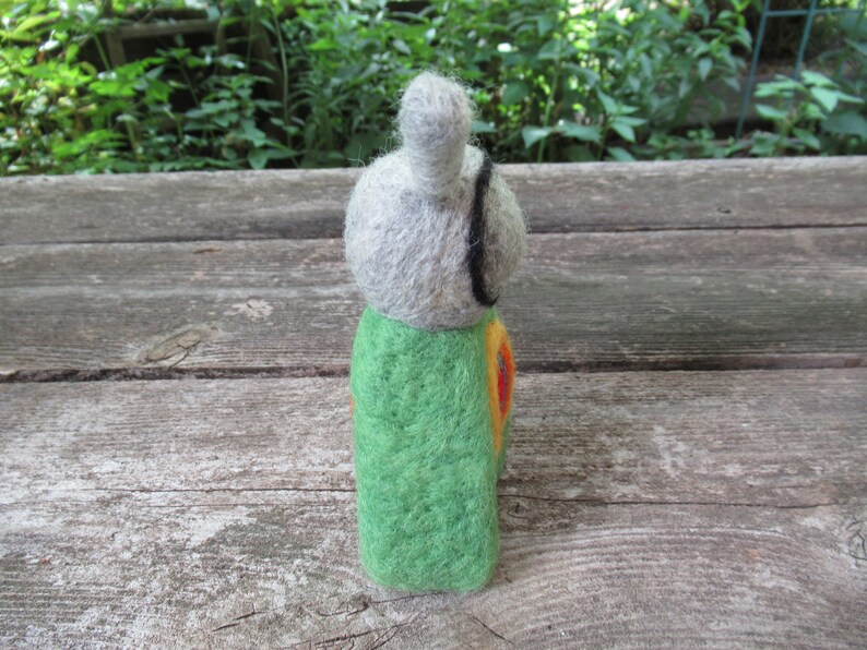 May include: A felt doll with a gray head, green body, and orange and yellow accents. The doll is standing on a wooden surface.