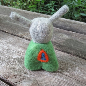 May include: A gray felt toy with long ears and a green felt body. The body has a red and blue felt design on the front.