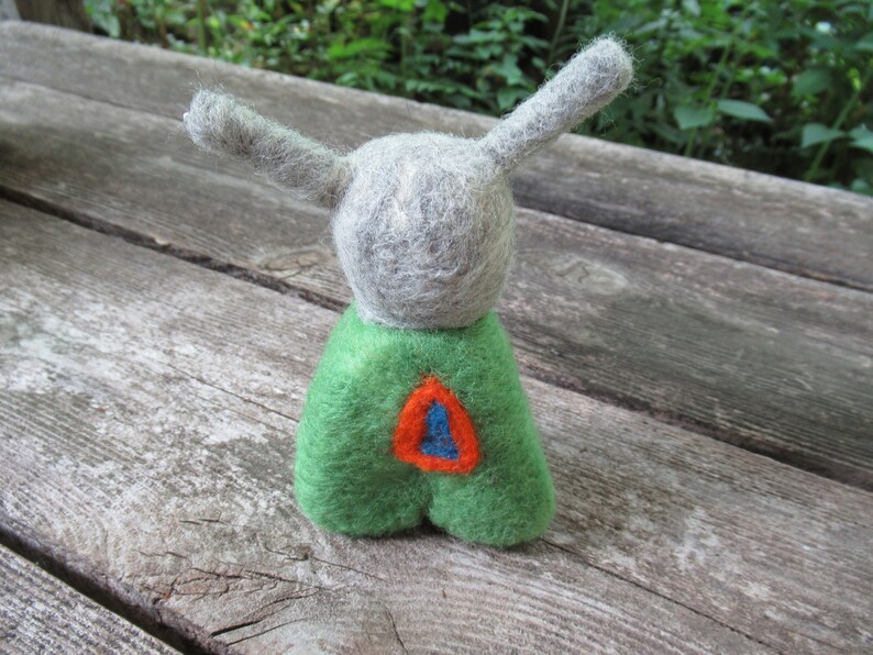 May include: A gray felt toy with long ears and a green felt body. The body has a red and blue felt design on the front.