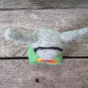 May include: A gray felt animal with long ears and a green, yellow, orange, and red felt body. The animal is sitting on a wooden surface.