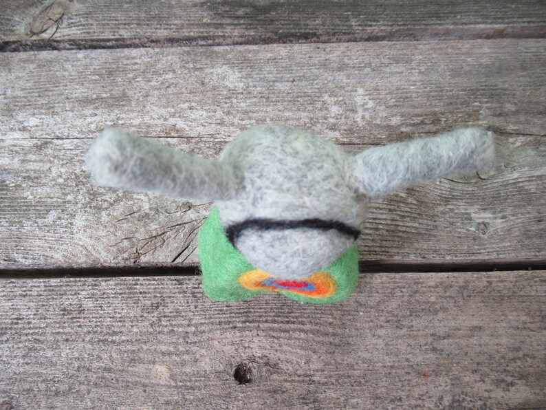 May include: A gray felt animal with long ears and a green, yellow, orange, and red felt body. The animal is sitting on a wooden surface.