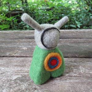 May include: A gray felt toy with long ears and a green felt body. The toy has a circular design on its chest with yellow, orange, blue, and red felt.