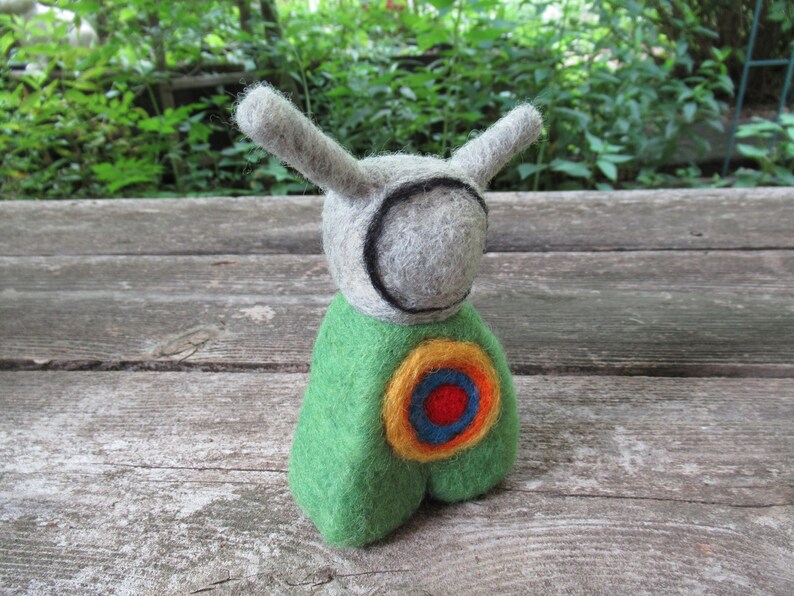 May include: A gray felt toy with long ears and a green felt body. The toy has a circular design on its chest with yellow, orange, blue, and red felt.