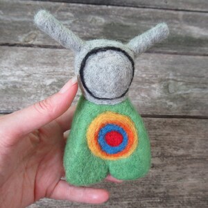 May include: A felt toy with a gray head and green body. The head has a black circle around it, and the body has a colorful target design with red, orange, yellow, and blue.
