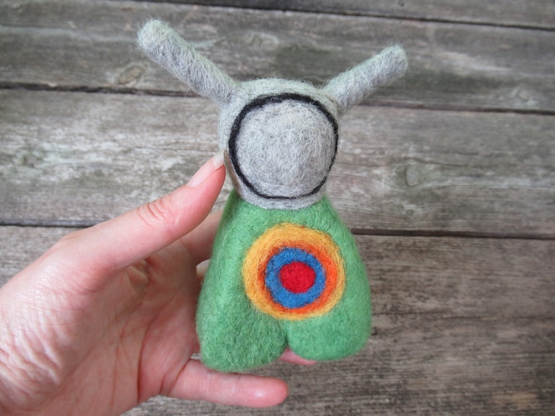 May include: A felt toy with a gray head and green body. The head has a black circle around it, and the body has a colorful target design with red, orange, yellow, and blue.