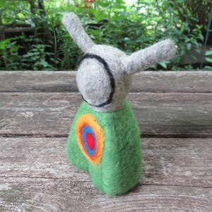 May include: A felt toy with a green body, gray head, and black eyes. The toy has a colorful circle design on its chest.