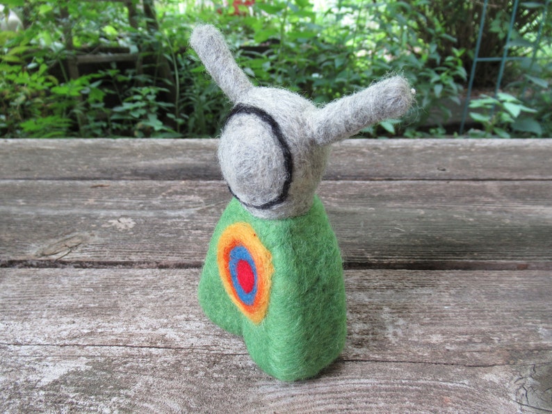 May include: A felt toy with a green body, gray head, and black eyes. The toy has a colorful circle design on its chest.