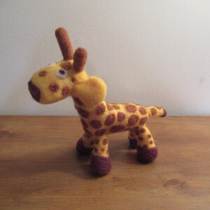 May include: A handmade felt giraffe toy. The giraffe is yellow with brown spots and accents. It has brown ears, a dark brown snout, and purple feet. The toy is standing on a wooden surface.