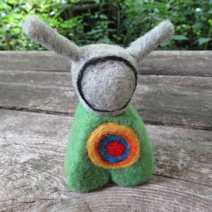 May include: A small felt toy with a gray head, green body, and a colorful target design on its chest. The toy has long, pointed ears.