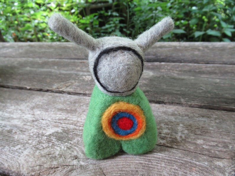 May include: A small felt toy with a gray head, green body, and a colorful target design on its chest. The toy has long, pointed ears.