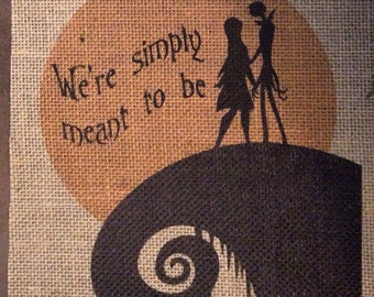 Op de afbeelding: Een wanddecoratie van jute stof met een silhouet van Jack Skellington en Sally uit de film The Nightmare Before Christmas. De achtergrond is een bruine cirkel met de tekst "We're simply meant to be".