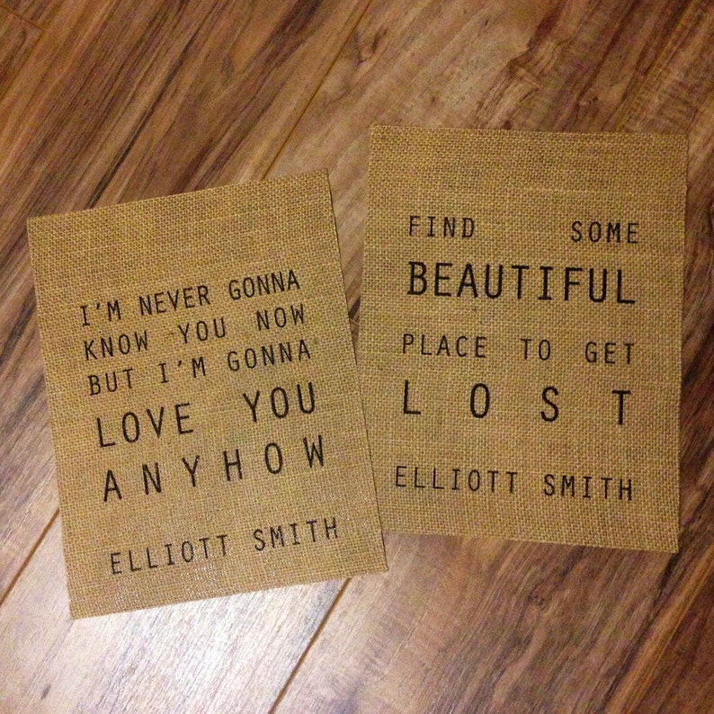 Op de afbeelding: Twee juteborden met zwarte tekst. Het eerste bord zegt "I'm never gonna know you now but I'm gonna love you anyhow Elliott Smith". Het tweede bord zegt "Find some beautiful place to get lost Elliott Smith".