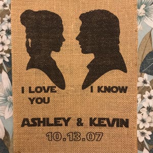 May include: A burlap wedding sign with black glitter silhouettes of a man and woman. The text reads "I love you" and "I know" with the couple's names "Ashley & Kevin" and wedding date "10.13.07".