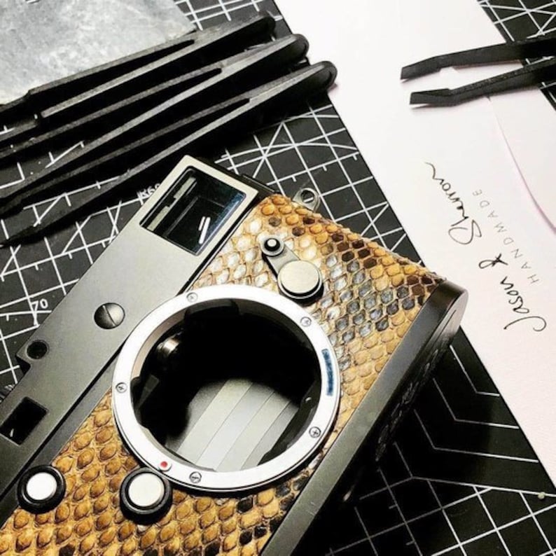 May include: A close-up of a camera with a snakeskin-textured body. The camera has a silver lens ring and black accents. The text "Jason & Shevrov Handmade" is visible on a white card.