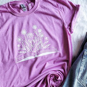 Peut inclure: Un t-shirt rose avec un imprim&eacute; blanc d'un livre ouvert avec des fleurs qui en sortent.