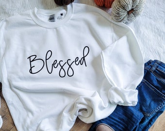 May include: White sweatshirt with the word "Blessed" written in black cursive lettering. The sweatshirt is laid out on a wooden surface with a pair of brown ankle boots, blue jeans, and three decorative pumpkins.