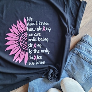 May include: A black t-shirt featuring a pink sunflower graphic on the left side and white text that reads: We don't know how strong we are until being strong is the only choice we have. The letter o in strong and choice is replaced by a pink ribbon.