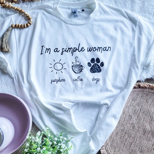 May include: White t-shirt with the text "I'm a simple woman" and three icons: a sun, a coffee cup, and a paw print. The text below the icons reads "sunshine, coffee, dogs".