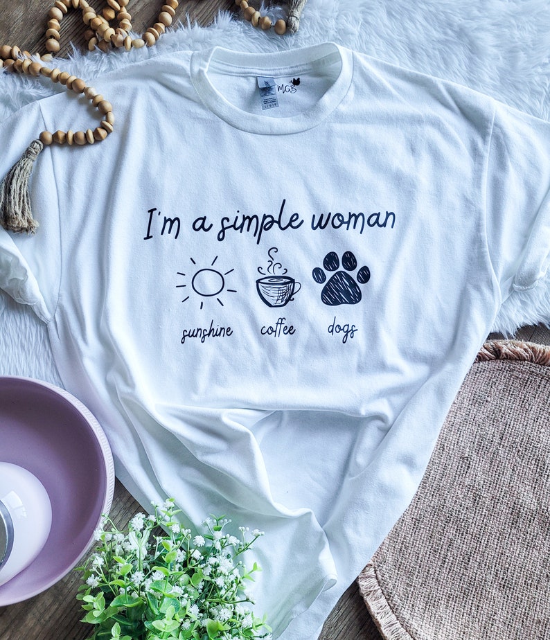 May include: White t-shirt with the text "I'm a simple woman" and three icons: a sun, a coffee cup, and a paw print. The text below the icons reads "sunshine, coffee, dogs".