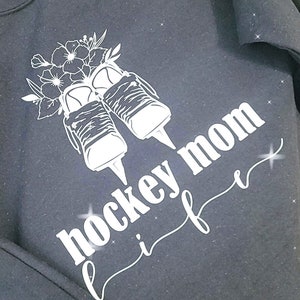 K&ouml;nnte beinhalten: Schwarzes Sweatshirt mit einem wei&szlig;en Design von Eishockeyschl&auml;gern mit Blumen und dem Text "Hockey Mom Life" in einer kursivschrift.