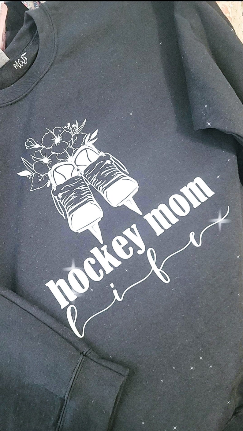 K&ouml;nnte beinhalten: Schwarzes Sweatshirt mit einem wei&szlig;en Design von Eishockeyschl&auml;gern mit Blumen und dem Text "Hockey Mom Life" in einer kursivschrift.