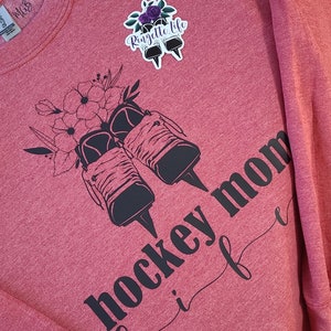 K&ouml;nnte beinhalten: Roter Pullover mit schwarzem Text, der "hockey mom life" lautet, und einer schwarz-wei&szlig;en Grafik von Eislaufschuhen mit Blumen. Der Text "Ringette Life" befindet sich auf einem Aufkleber auf dem Pullover.