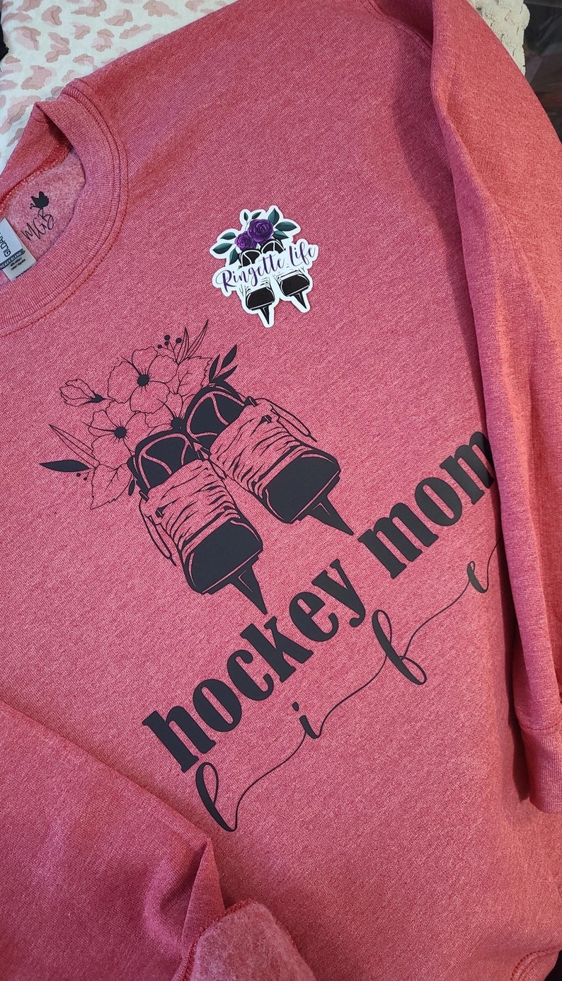 K&ouml;nnte beinhalten: Roter Pullover mit schwarzem Text, der "hockey mom life" lautet, und einer schwarz-wei&szlig;en Grafik von Eislaufschuhen mit Blumen. Der Text "Ringette Life" befindet sich auf einem Aufkleber auf dem Pullover.