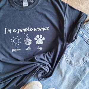 May include: A black t-shirt with the text "I'm a simple woman" and three icons: a sun, a coffee cup, and a paw print. The icons are labeled "sunshine", "coffee", and "dogs".