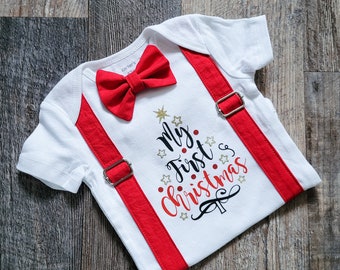 May include: White baby bodysuit with red suspenders and a matching bow tie. The bodysuit features the text "My First Christmas" in red and black, with a Christmas tree design.