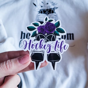 K&ouml;nnte beinhalten: Ein wei&szlig;er Aufkleber mit einer schwarz-wei&szlig;en Illustration von Schlittschuhen mit lila Blumen und dem Text "Hockey Life".