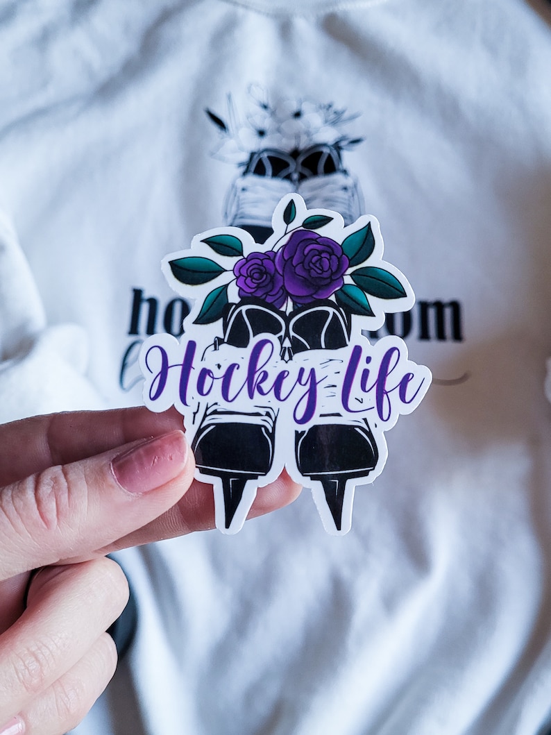 K&ouml;nnte beinhalten: Ein wei&szlig;er Aufkleber mit einer schwarz-wei&szlig;en Illustration von Schlittschuhen mit lila Blumen und dem Text "Hockey Life".