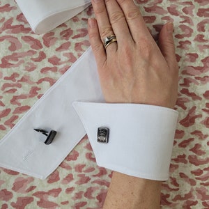 May include: White dress shirt cuffs with black cufflinks. The cufflinks have a rectangular design with a raised pattern. The shirt cuffs are folded back to show the cufflinks.