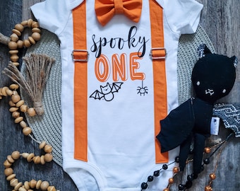 Peut inclure: Body bébé blanc avec bretelles et nœud papillon orange. Le body porte l'inscription "spooky ONE" avec des graphiques de chauve-souris et d'araignée. Une peluche chat noir et une guirlande de perles sont également dans l'image. Tenue de bébé sur le thème d'Halloween.