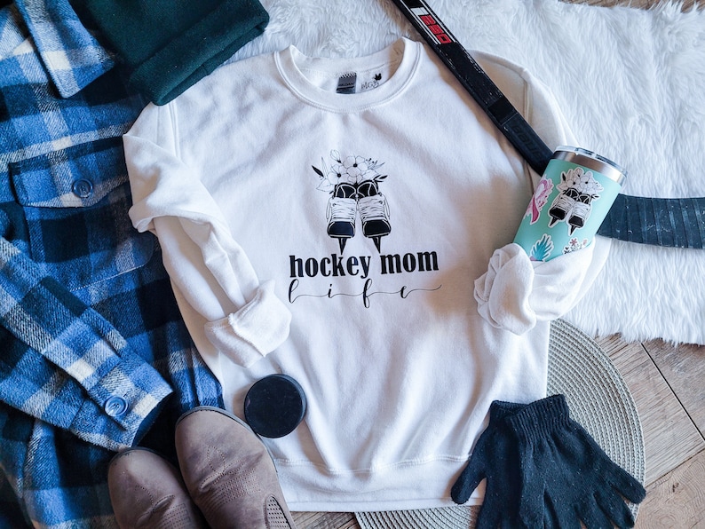 K&ouml;nnte beinhalten: Wei&szlig;es Sweatshirt mit schwarzem Text, der "hockey mom life" lautet, und einer Grafik von Eishockeyschuhen mit Blumen. Das Sweatshirt liegt auf einer wei&szlig;en, flauschigen Oberfl&auml;che mit einem Hockeyschl&auml;ger, einem blau-wei&szlig; karierten Hemd, braunen Stiefeln, einer schwarzen Scheibe und einem schwarz-wei&szlig; gemusterten Becher.
