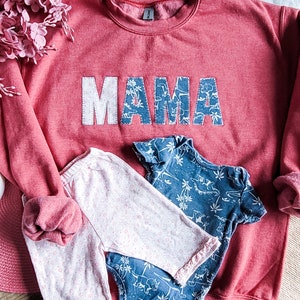 May include: A red sweatshirt with the word "MAMA" in white and blue patchwork letters. The image also shows baby clothes: pink floral trousers and a blue dinosaur print babygrow.