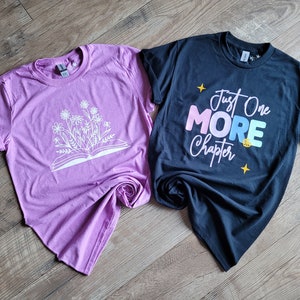 Peut inclure: Deux t-shirts, un rose et un noir. Le t-shirt rose a un motif blanc d'un livre ouvert avec des fleurs qui en sortent. Le t-shirt noir a un motif blanc qui dit "Just One More Chapter" avec le mot "More" en couleurs arc-en-ciel.