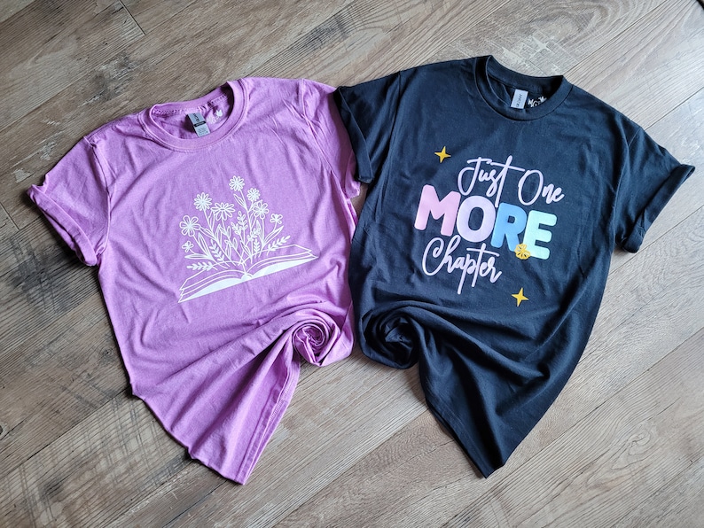 Peut inclure: Deux t-shirts, un rose et un noir. Le t-shirt rose a un motif blanc d'un livre ouvert avec des fleurs qui en sortent. Le t-shirt noir a un motif blanc qui dit "Just One More Chapter" avec le mot "More" en couleurs arc-en-ciel.