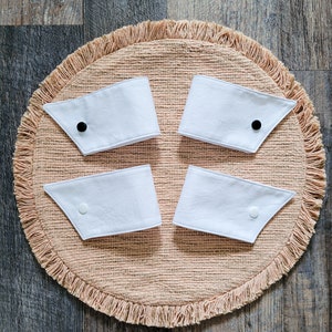 May include: Four white fabric collar cuffs with black buttons on a beige fringed round rug.